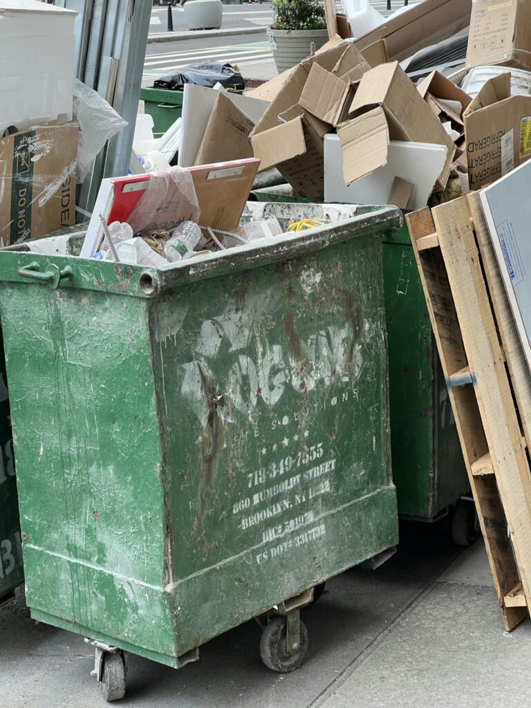 Green dumpster on city street filled with cardboard and debris, highlighting urban waste management.